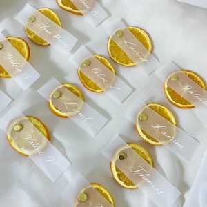 Puede incluir: Tarjetas de lugar para una boda o evento, con rodajas de naranja secas con nombres escritos en tarjetas de papel translúcido. Las tarjetas están sujetas a las rodajas de naranja con alfileres dorados.