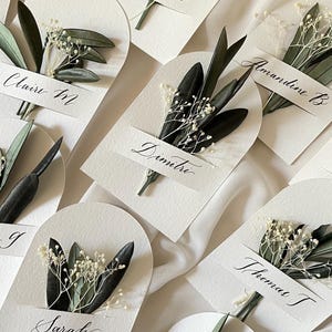 Puede incluir: Tarjetas de lugar blancas con nombres en caligrafía y pequeños ramos de flores blancas secas y hojas verdes.