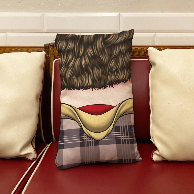 Op de afbeelding: Een decoratief kussen met een geruit patroon en een cartoonafbeelding van de rug van een persoon met bruin haar, een rode kraag en een geel shirt.