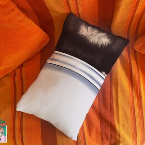May include: A rectangular pillow with a black and white graphic design, resting on a striped orange and red surface. The pillow has a white lower section and a black upper section with white details. Two orange pillows are also visible.