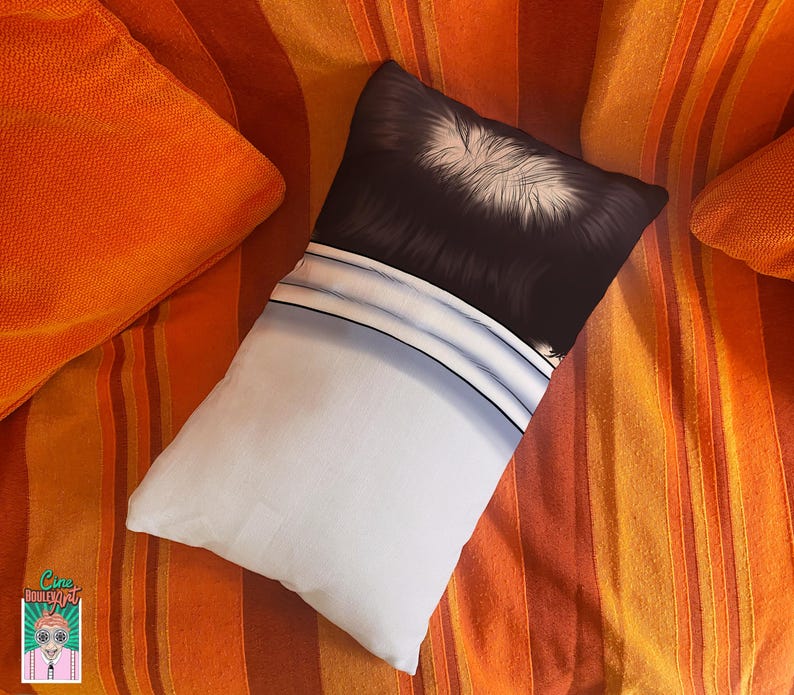 May include: A rectangular pillow with a black and white graphic design, resting on a striped orange and red surface. The pillow has a white lower section and a black upper section with white details. Two orange pillows are also visible.