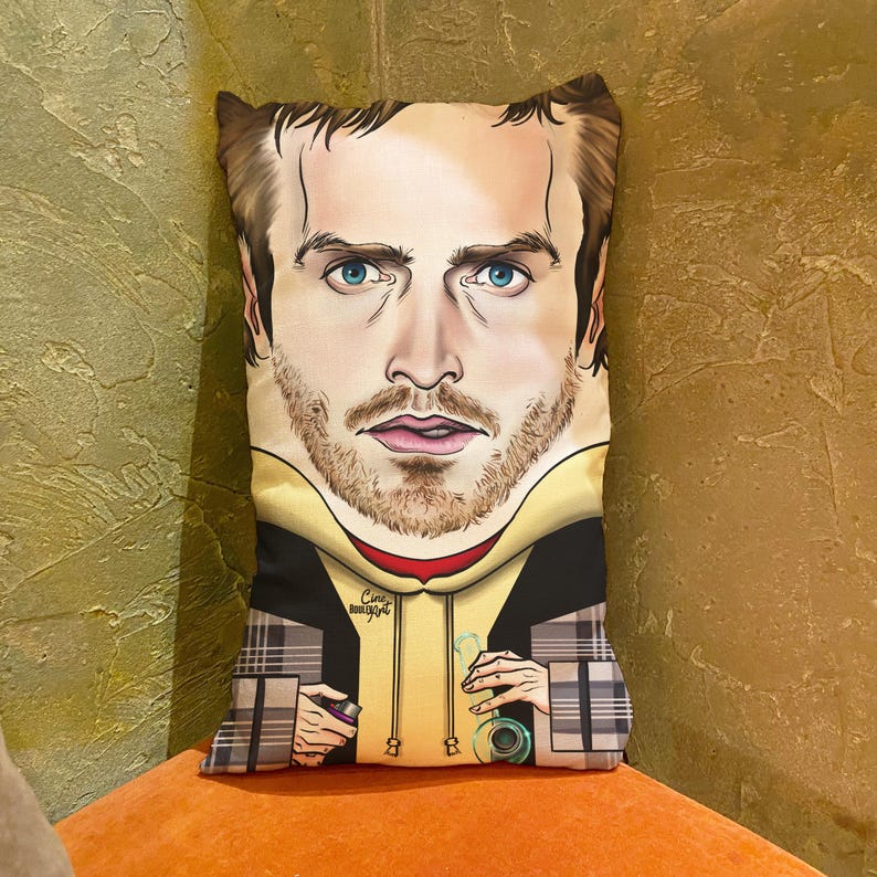 Op de afbeelding: Een decoratief kussen met een cartoonillustratie van een man met een baard en een gele hoodie. De man houdt een glazen pijp in de ene hand en een spuitbus in de andere. Het kussen heeft een geruit patroon op de onderste helft.
