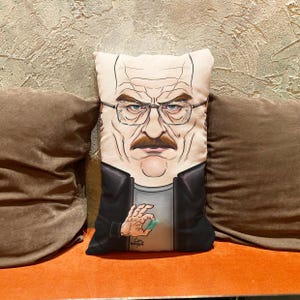 Op de afbeelding: Een decoratief kussen met een cartoonillustratie van Walter White uit de tv-serie Breaking Bad. Het kussen is wit met een zwarte achtergrond en toont Walter White die een blauwe kristal in zijn hand houdt. De tekst "Cine" staat in de rechterbenedenhoek van het kussen.