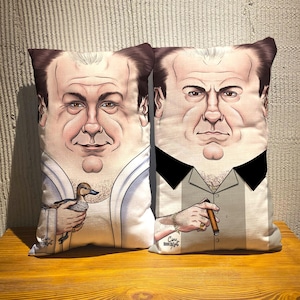 May include: Two decorative throw pillows with a caricature of a man's face. One pillow features a man holding a duck, the other a man holding a cigar. Both pillows have a light background and are on a wooden surface.