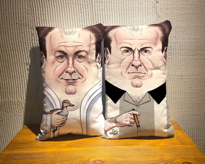 May include: Two decorative throw pillows with a caricature of a man's face. One pillow features a man holding a duck, the other a man holding a cigar. Both pillows have a light background and are on a wooden surface.