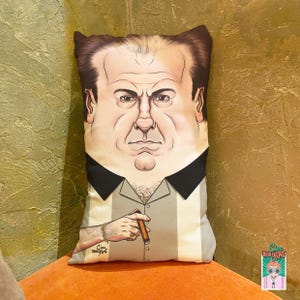 May include: A decorative pillow featuring a stylized illustration of a man with a cigar. The pillow has a light gray and white striped shirt with a black collar. The illustration includes a gold bracelet and ring. The pillow is rectangular.
