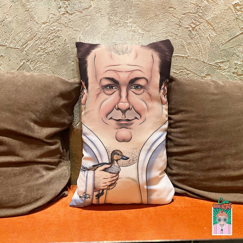 May include: A rectangular decorative pillow featuring a caricature of a man holding a duck. The pillow has a white background with a portrait of a man with dark hair and a light complexion. The duck is brown and gray.