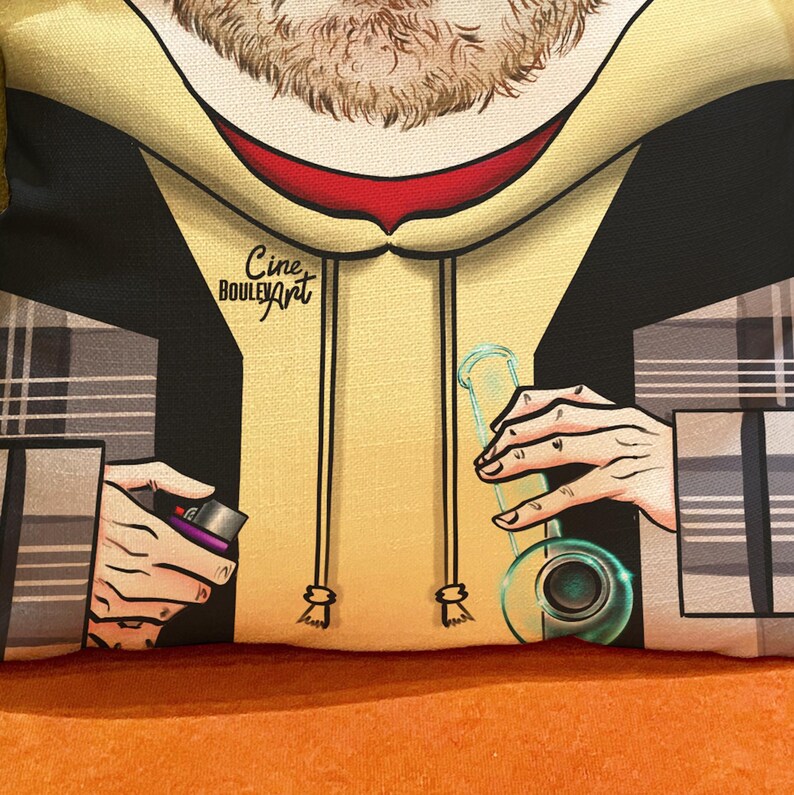 Op de afbeelding: Een gele hoodie met een zwart-wit geruit patroon op de mouwen. De hoodie heeft een rode kraag en de tekst "Cine Bouley Art" staat op de voorkant. De persoon op de afbeelding houdt een glazen pijp en een aansteker vast.