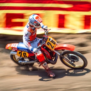 Peut inclure: Un pilote de motocross portant un casque bleu et blanc avec une combinaison de course rouge et blanche, conduisant une moto de cross rouge et blanche avec le numéro 26 sur le côté. Le pilote est en mouvement, et l'arrière-plan est flou.