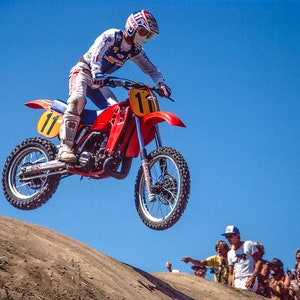 Peut inclure: Une moto de motocross rouge et blanche avec le numéro 11 sur le côté, en plein vol, avec un pilote portant un casque et un équipement de course. La moto est sur fond de ciel bleu avec une colline de terre au premier plan et des spectateurs en arrière-plan.