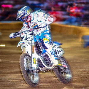 May include: A motocross racer wearing a white, blue, and red racing suit with the number 25 on their jersey, riding a white and blue dirt bike on a dirt track.