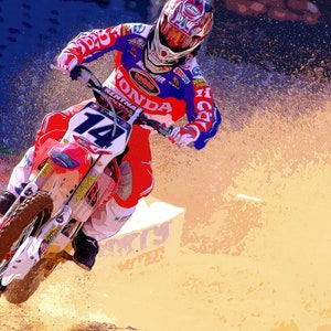 Może przedstawiać: Motocyklista w czerwono-biało-niebieskim kombinezonie wyścigowym z numerem "14" na motocyklu, jadący po torze ziemnym z chmurą kurzu za sobą.