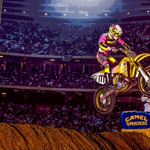 Puede incluir: Un piloto de motocross con un jersey rosa y amarillo salta sobre una rampa con un cartel de "Camel Supercross". El piloto lleva casco y gafas. El fondo muestra un gran estadio lleno de espectadores.