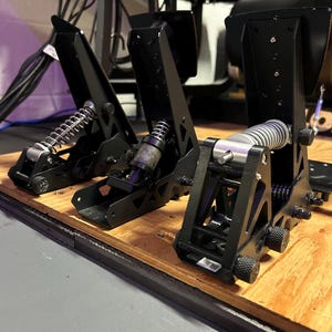 May include: Close-up of three black racing pedals with silver springs and adjustable knobs. The pedals are mounted on a wooden base, showcasing a high-performance racing simulator setup. The design emphasizes precision and control.