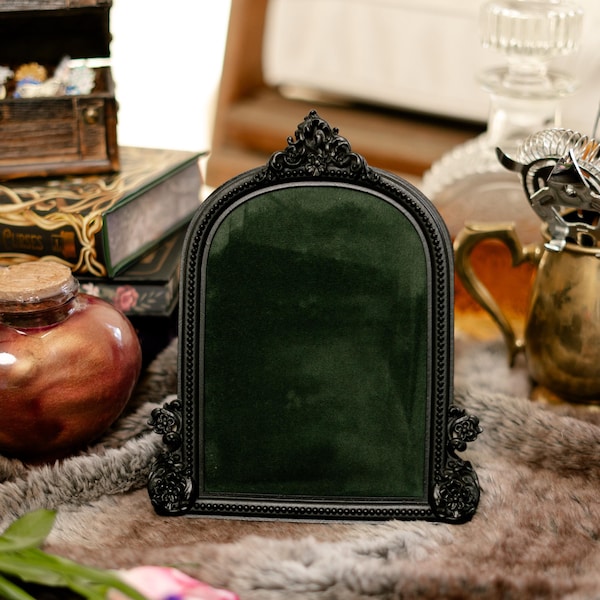 May include: A black ornate picture frame with a dark green velvet insert. The frame is sitting on a brown faux fur surface.