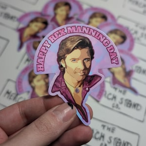 May include: A pink and blue sticker with a cartoon portrait of a man with brown hair and a pink shirt. The sticker has the text "Happy Rex Manning Day" in a pink and white font.