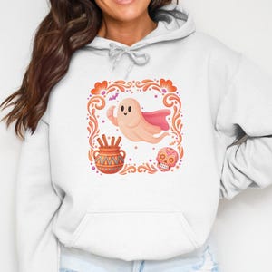 May include: A light gray hooded sweatshirt featuring a central graphic print of a peach-colored ghost wearing a pink cape, a brown patterned pot with sticks, and a pink sugar skull, all framed by orange floral flourishes and small bats.