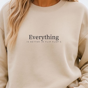 Könnte beinhalten: Beiges Sweatshirt mit dem schwarzen Text "Everything is better in flip flops". Das Sweatshirt hat eine neutrale Farbe und einen Rundhalsausschnitt. Das Model trägt eine blaue Jeans.