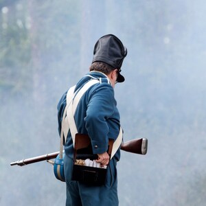 May include: A man in a blue uniform with white straps and a black hat is holding a rifle. He is wearing a leather pouch with white paper cartridges on his belt.