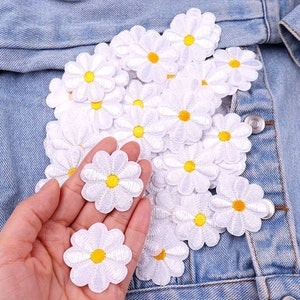 May include: A collection of white felt daisy flower patches with yellow centres. The patches are arranged in a pile on a blue denim jacket.