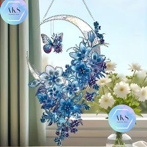 May include: A decorative crescent moon-shaped wall hanging with blue floral and butterfly accents. The piece features a silver chain for hanging and is adorned with various shades of blue flowers and a butterfly pendant. The design is detailed with sparkling embellishments.