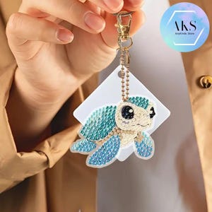 May include: A bejeweled turtle keychain with a gold-colored clasp and chain. The turtle features blue and teal gems on its shell and flippers, with a cream-colored body and black eyes. A white tag is attached. The keychain is from the AnyKinds Store.