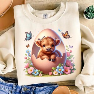 May include: A cream-coloured sweatshirt featuring a graphic of a brown and white Highland calf inside a pink egg surrounded by colourful flowers and butterflies.