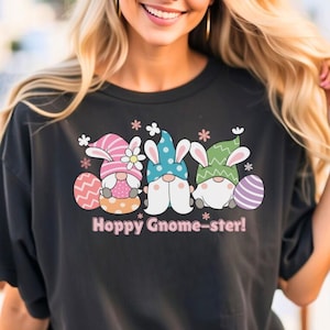 以下が含まれることがあります： イースターバニーの耳をつけた3人のノームがイースターエッグを持っているグラフィックがプリントされた黒いTシャツ。ノームの下には「Hoppy Gnome-ster!」という文字がプリントされています。