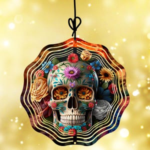 May include: A Day of the Dead-themed ornament featuring a decorated skull with floral accents. The skull is adorned with colorful flowers and intricate patterns, suspended by a black string against a gold background.