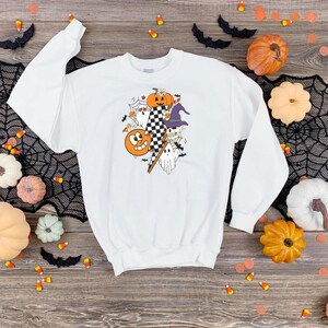 May include: White sweatshirt featuring a Halloween-themed graphic. The design includes a smiling jack-o'-lantern, pumpkins, ghosts, a witch's hat, and a checkered flag. The sweatshirt is surrounded by Halloween decorations, including pumpkins and candy corn.