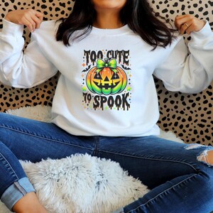 May include: White long-sleeve sweatshirt with a colorful pumpkin graphic. The pumpkin has a green bow and the text "TOO CUTE TO SPOOK". The design is in orange, green, yellow, and black. The sweatshirt is worn with blue jeans.