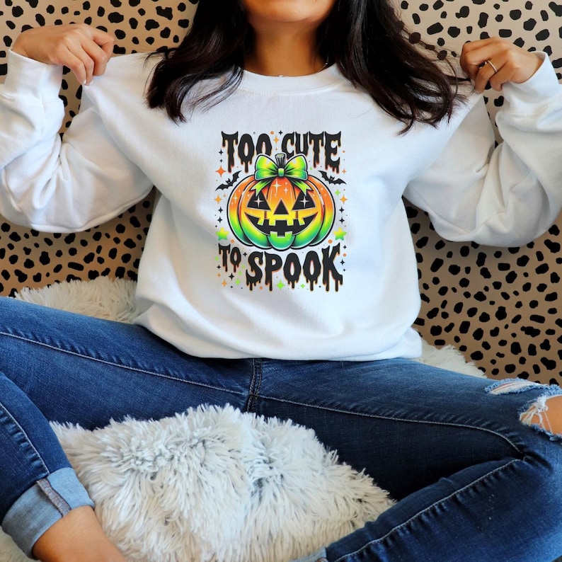 May include: White long-sleeve sweatshirt with a colorful pumpkin graphic. The pumpkin has a green bow and the text "TOO CUTE TO SPOOK". The design is in orange, green, yellow, and black. The sweatshirt is worn with blue jeans.