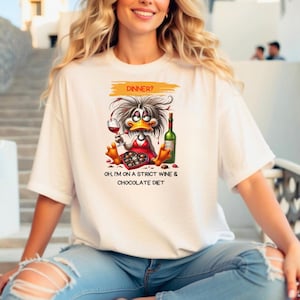 May include: White t-shirt featuring a cartoon duck enjoying wine and chocolate. The duck is wearing a red dress and holding a wine glass. Text on the shirt reads "Dinner?" and "Oh, I'm on a strict wine & chocolate diet."