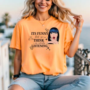 Puede incluir: Camiseta naranja de manga corta con el texto "ITS FUNNY that you THINK I am actually LISTENING" y una ilustraci&oacute;n de estilo pop art de una mujer con pelo azul. La camiseta es de material suave.