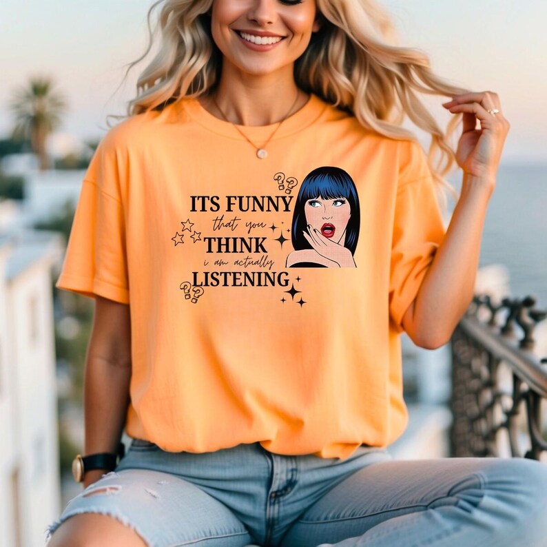 Puede incluir: Camiseta naranja de manga corta con el texto "ITS FUNNY that you THINK I am actually LISTENING" y una ilustraci&oacute;n de estilo pop art de una mujer con pelo azul. La camiseta es de material suave.