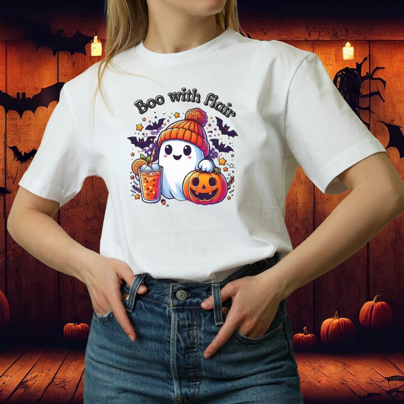 May include: White t-shirt with a Halloween-themed graphic. The design features a cartoon ghost wearing an orange beanie, holding a pumpkin and a drink. The text "Boo with flair" is above the ghost.