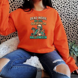 May include: Orange sweatshirt with the text "DEAD INSIDE BUT STILL QUEUED FOR HALLOWEEN". The graphic features a skeleton wearing headphones and holding a game controller and pizza. The sweatshirt is a long-sleeved crew neck.