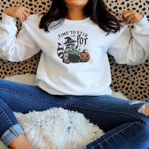 May include: White long-sleeve sweatshirt with a Halloween-themed graphic. The graphic features a raccoon wearing a witch hat, stirring a cauldron with the text "TIME TO STIR THE POT" and two pumpkins. The raccoon is gray, black, and white.