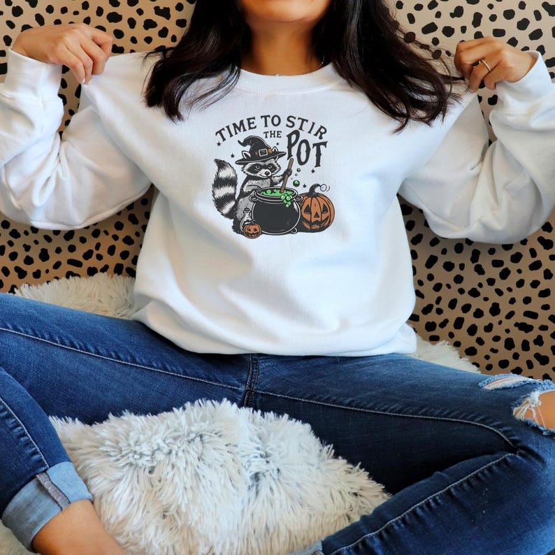 May include: White long-sleeve sweatshirt with a Halloween-themed graphic. The graphic features a raccoon wearing a witch hat, stirring a cauldron with the text "TIME TO STIR THE POT" and two pumpkins. The raccoon is gray, black, and white.