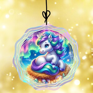 May include: A whimsical ornament featuring a white unicorn with a colorful mane and tail, nestled in a woven nest. The background showcases a vibrant aurora borealis scene, framed by a wavy, light purple border. A black string is attached for hanging.