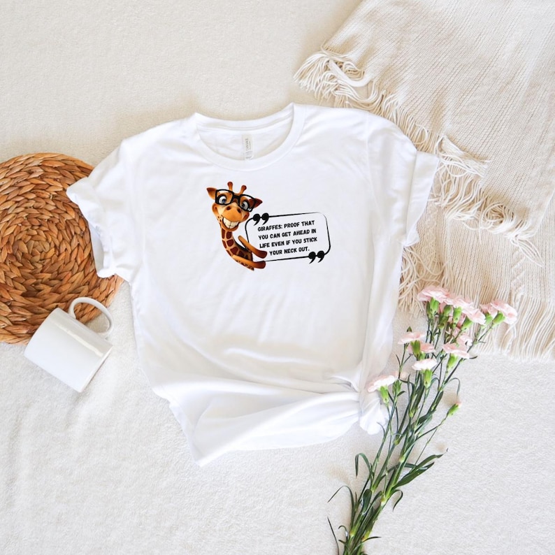 May include: White t-shirt featuring a cartoon giraffe wearing glasses. The giraffe is smiling and has a speech bubble with the text: "Giraffes: Proof that you can get ahead in life even if you stick your neck out."