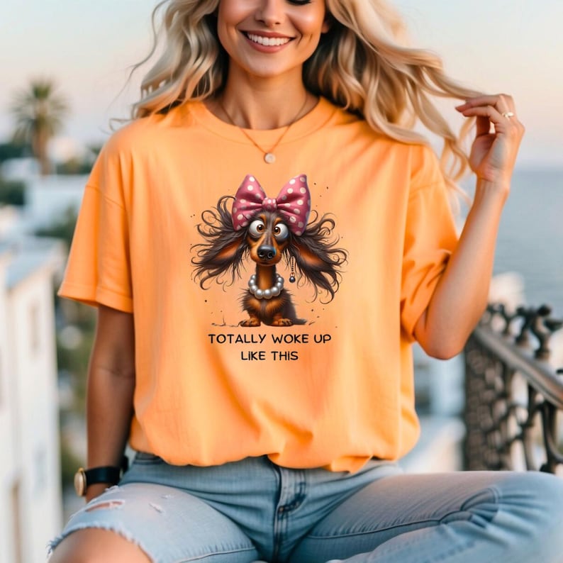May include: Orange short-sleeved t-shirt featuring a cartoon dachshund with a pink polka-dot bow, pearl necklace, and the text "TOTALLY WOKE UP LIKE THIS". The dog has long, flowing hair. The shirt is made of a soft material.