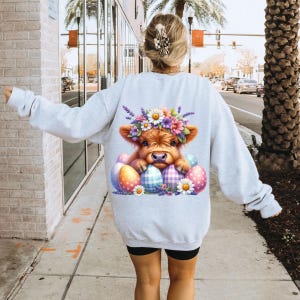 May include: A gray sweatshirt with a graphic of a brown and white Highland cow wearing a flower crown and surrounded by colorful Easter eggs.