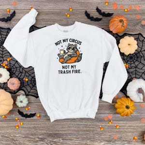 May include: White sweatshirt with a graphic of a raccoon wearing sunglasses and holding a mug. The text on the sweatshirt reads "NOT MY CIRCUS NOT MY TRASH FIRE." The sweatshirt is surrounded by Halloween decorations.