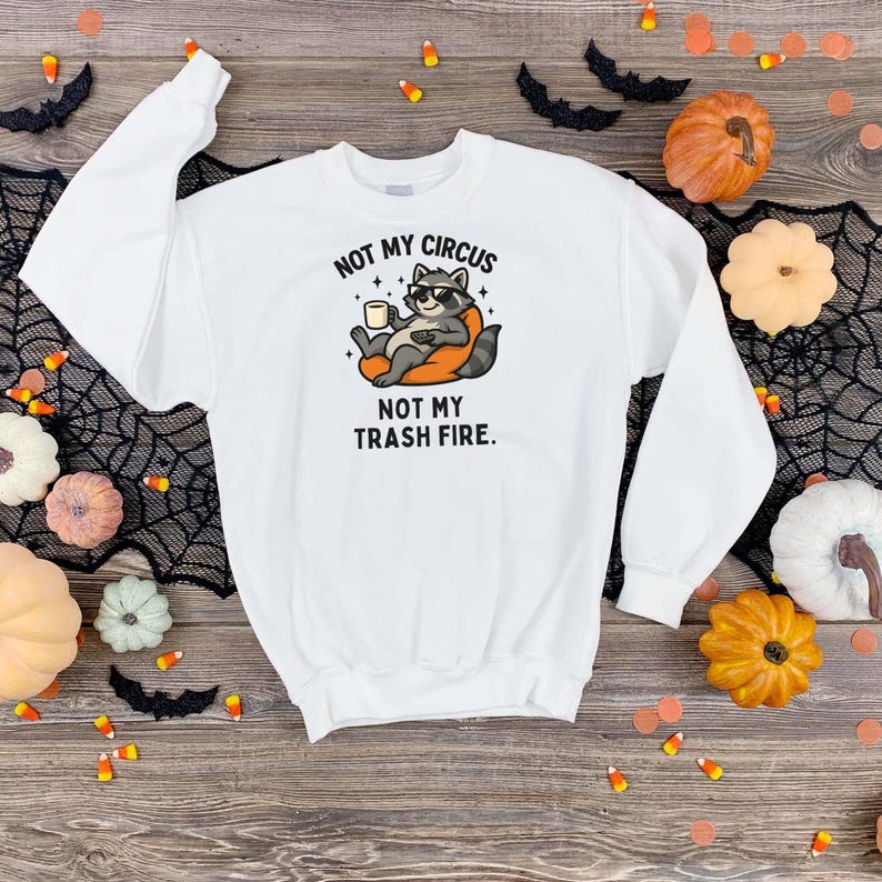 May include: White sweatshirt with a graphic of a raccoon wearing sunglasses and holding a mug. The text on the sweatshirt reads "NOT MY CIRCUS NOT MY TRASH FIRE." The sweatshirt is surrounded by Halloween decorations.