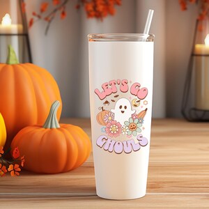 May include: A white tumbler with a straw featuring a Halloween-themed design. The design includes a friendly ghost, pumpkins, flowers, and the text "Let's Go Ghouls" in pastel colors. The tumbler is set against a background of pumpkins and candles.