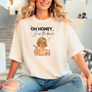 Puede incluir: Camiseta blanca con la frase "OH HONEY I am the drama" y un gr&aacute;fico de una mujer con cabello rubio, gafas rojas y l&aacute;piz labial. La mujer lleva un top rosa. La camiseta es de un material suave.