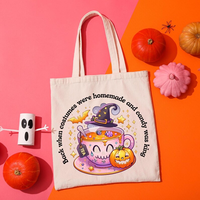 May include: A cream-colored tote bag with a Halloween-themed design. The graphic features a smiling teacup wearing a witch hat, a pumpkin, and the text "Back when costumes were homemade and candy was king."