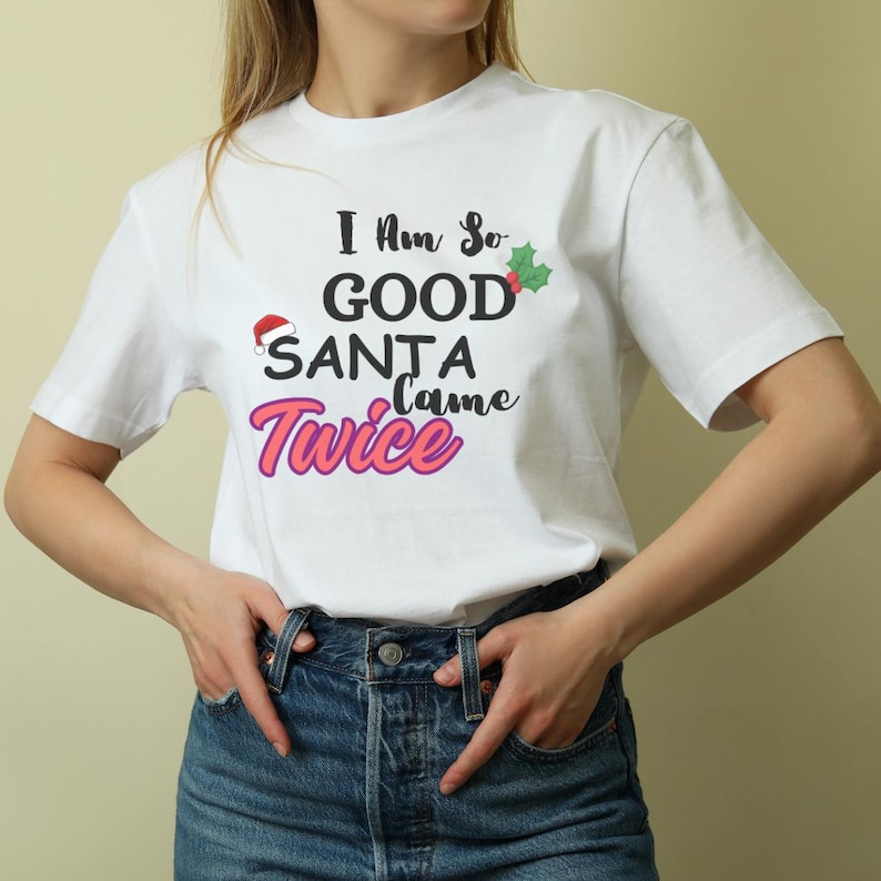 May include: White t-shirt with the text "I am so good Santa came twice" in black and pink lettering. A small red Santa hat and a green holly sprig are included in the design.