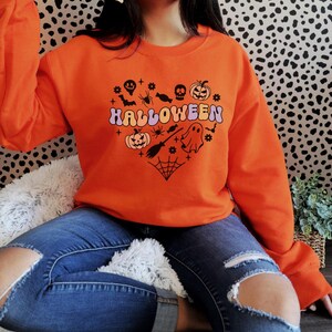 May include: Orange Halloween sweatshirt with a heart-shaped graphic. The graphic includes a pumpkin, spider, bat, ghost, and the word "HALLOWEEN" in a retro font. The sweatshirt is made of a soft material and is perfect for the fall season.